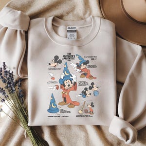 Puede incluir: Una sudadera de cuello redondo en color beige con un estampado gr&aacute;fico de Mickey Mouse como el aprendiz de brujo de Fantas&iacute;a. El dise&ntilde;o incluye varias ilustraciones del personaje, texto descriptivo y etiquetas t&eacute;cnicas en tinta negra y azul.