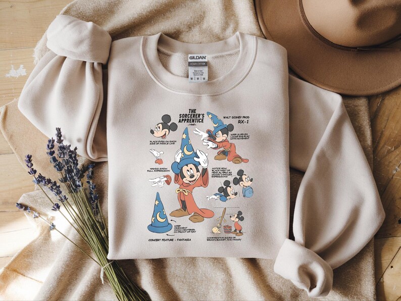Puede incluir: Una sudadera de cuello redondo en color beige con un estampado gr&aacute;fico de Mickey Mouse como el aprendiz de brujo de Fantas&iacute;a. El dise&ntilde;o incluye varias ilustraciones del personaje, texto descriptivo y etiquetas t&eacute;cnicas en tinta negra y azul.
