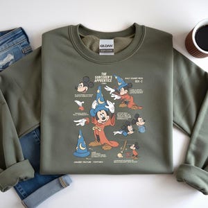 Puede incluir: Una sudadera de cuello redondo color verde oliva con un estampado gr&aacute;fico de Mickey Mouse como el aprendiz de brujo. El dise&ntilde;o incluye varias ilustraciones del personaje, un sombrero de mago azul y texto descriptivo sobre la pel&iacute;cula Fantas&iacute;a de 1940.