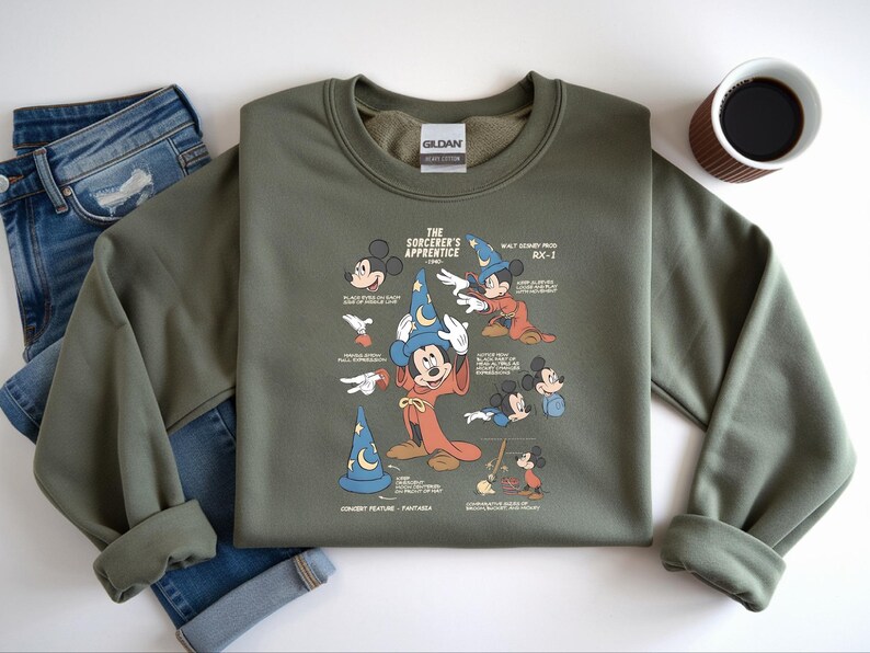 Puede incluir: Una sudadera de cuello redondo color verde oliva con un estampado gr&aacute;fico de Mickey Mouse como el aprendiz de brujo. El dise&ntilde;o incluye varias ilustraciones del personaje, un sombrero de mago azul y texto descriptivo sobre la pel&iacute;cula Fantas&iacute;a de 1940.
