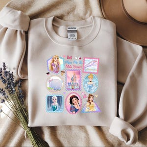 May include: Beige crewneck sweatshirt featuring colorful postage stamp-style graphics of Disney princesses, castles, and other themed images. The text "Meet Me at the Sweet" is also included. The sweatshirt is made of a soft material.