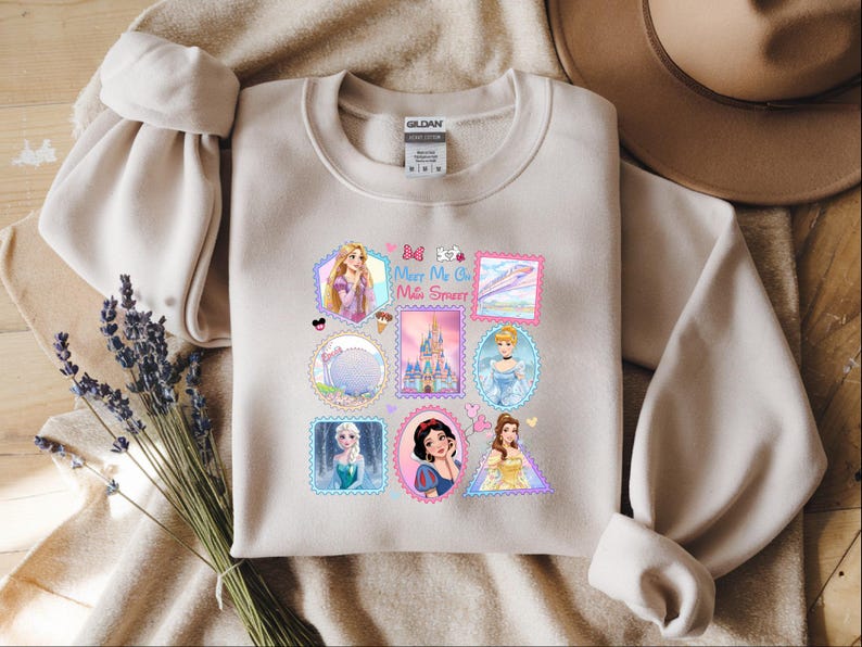 May include: Beige crewneck sweatshirt featuring colorful postage stamp-style graphics of Disney princesses, castles, and other themed images. The text "Meet Me at the Sweet" is also included. The sweatshirt is made of a soft material.
