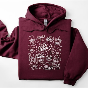 May include: A maroon hooded sweatshirt featuring a white graphic print of various Dr Pepper themed icons, including a soda can, a bottle, a six-pack, a cowboy hat, a foam finger, cherries, bows, hearts, stars, and the text Dr Pepper and 23.