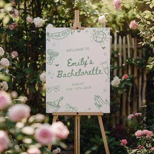 May include: A white sign on a wooden easel with green text and illustrations. The sign reads "Welcome to Emily's Bachelorette" with the date "August 4th-6th, 2026". The sign is surrounded by a garden with pink and white roses.