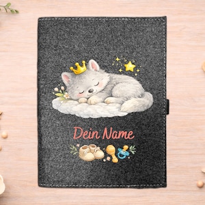 Könnte beinhalten: Eine dunkelgraue Babybuchhülle aus Filz mit einem schlafenden Wolfswelpen mit goldener Krone. Auf der Hülle steht der Text "Dein Name" in Rosa, zusammen mit Illustrationen von Babyschuhen, einer Rassel und einem Schnuller. Sterne und Blumen sind ebenfalls enthalten.