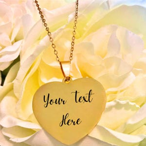 May include: A gold-colored heart-shaped pendant necklace with the words "Your text Here" in black script. The necklace hangs from a delicate gold chain. The background is a soft focus of yellow and white flowers.
