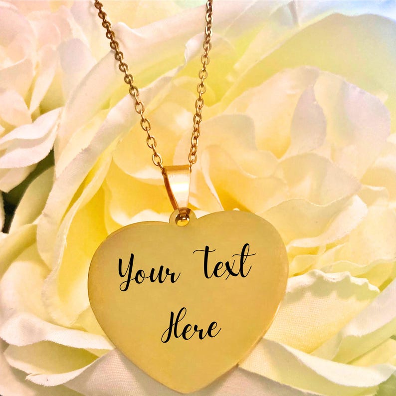 May include: A gold-colored heart-shaped pendant necklace with the words "Your text Here" in black script. The necklace hangs from a delicate gold chain. The background is a soft focus of yellow and white flowers.