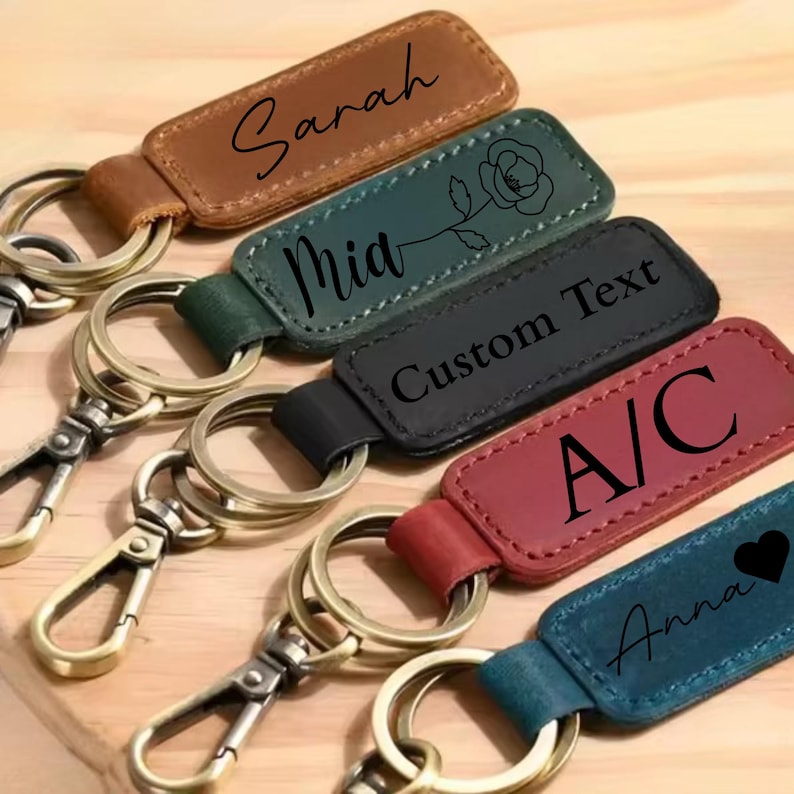 May include: A collection of personalized leather keychains in various colors, including brown, green, black, red, and blue. Each keychain features a metal ring and clasp, with custom text or designs such as names, initials, and a floral motif.