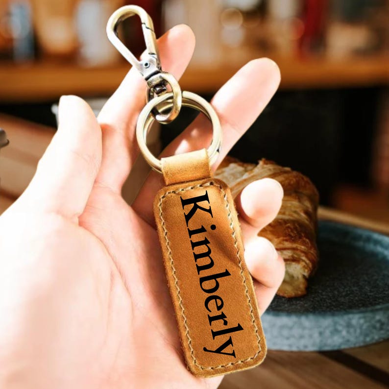 May include: A tan leather keychain with the name "Kimberly" in black lettering. The keychain has a metal clasp and a metal ring. The background is blurred, but includes a croissant.