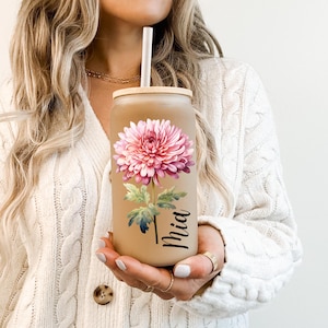 May include: A beige glass tumbler with a bamboo lid and straw, featuring a large pink chrysanthemum flower design. The name "Mia" is printed vertically on the glass. The tumbler is held by a person wearing a white sweater.