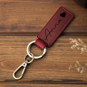 May include: A burgundy leather keychain with the name "Anna" in cursive and a black heart symbol. The keychain has a silver metal ring and a clasp. The background is a wooden surface.