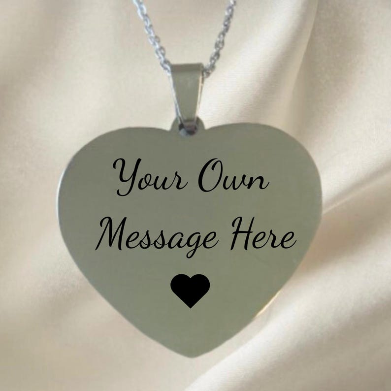May include: A silver heart-shaped pendant necklace. The pendant has the text "Your Own Message Here" in black script, with a small black heart at the bottom. The necklace chain is also silver.