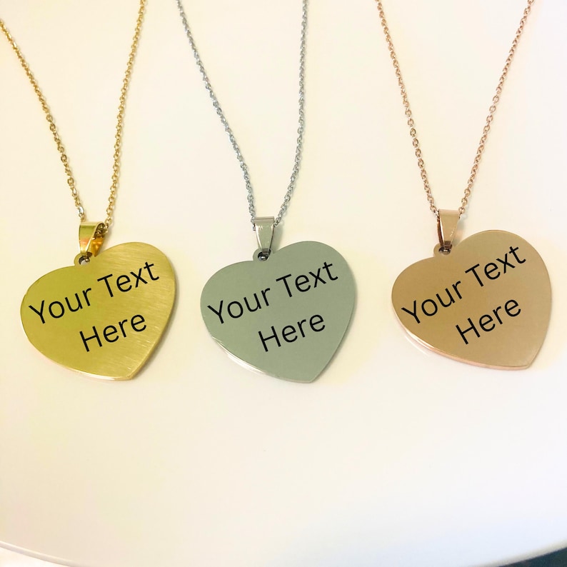 May include: Three heart-shaped pendant necklaces in gold, silver, and rose gold. Each pendant has a chain and the text "Your Text Here" in black. The pendants are on a white surface.
