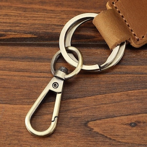 May include: Close-up of a bronze-colored keychain with a swivel clasp and a large key ring. The keychain features a leather strap in a medium brown color, attached to the key ring. The background is a wooden surface.