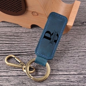 May include: A teal leather keychain with a gold-colored clasp and ring. The keychain is engraved with the letter "B" and the name "Brittney". The keychain is displayed on a wooden surface.
