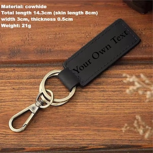 May include: Black cowhide keychain with metal hardware. The keychain has the text "Your Own Text" engraved on it. Total length is 5.6 inches, width is 1.2 inches, and thickness is 0.2 inches. The keychain weighs 0.7 ounces.