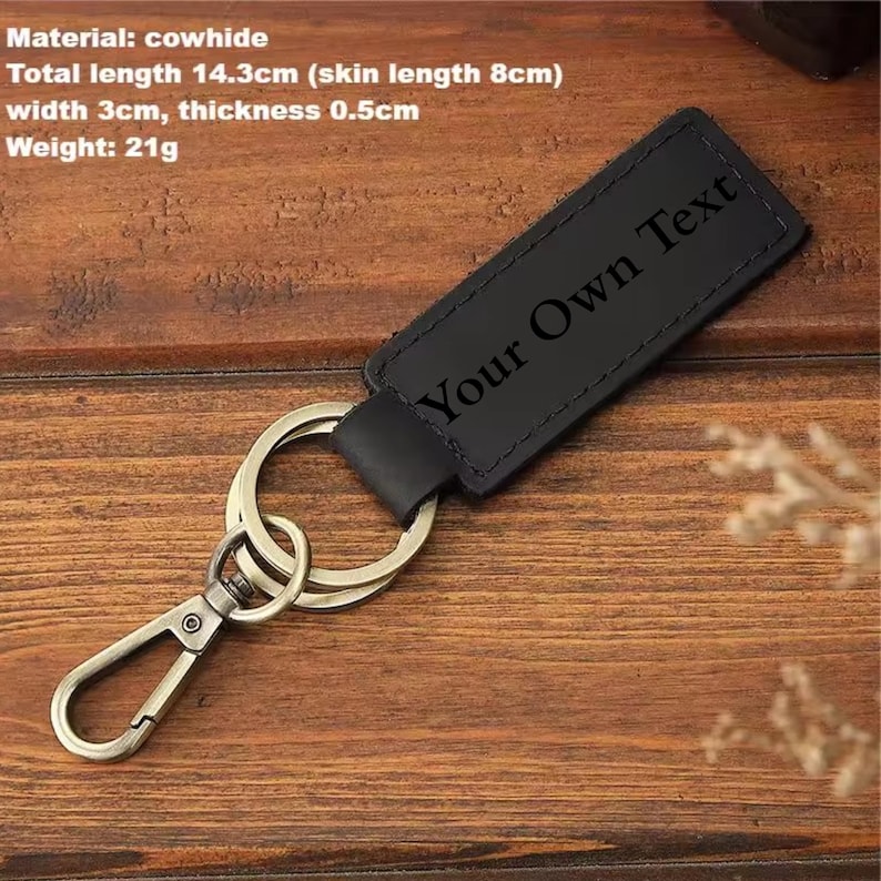 May include: Black cowhide keychain with metal hardware. The keychain has the text "Your Own Text" engraved on it. Total length is 5.6 inches, width is 1.2 inches, and thickness is 0.2 inches. The keychain weighs 0.7 ounces.