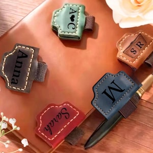 May include: Five leather pen holders in green, brown, dark gray, blue, and red, each featuring an elastic band and personalized text or initials including A heart C, SR, Anna, M, and Sarah, attached to a brown leather notebook.