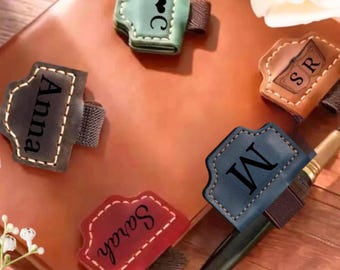 May include: Five leather pen holders in green, brown, dark gray, red, and blue, each featuring an elastic band and personalized text or initials including A heart C, SR, Anna, M, and Sarah, attached to a brown leather notebook.