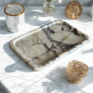 Może przedstawiać: Prostokątna marmurowa taca z kremowym i ciemnobrązowym wzorem żyłkowym. Taca znajduje się na marmurowej powierzchni, z marmurowym kubkiem ze złotą obwódką, małym szklanym wazonem z białymi kwiatami i złotą dekoracyjną kulą.