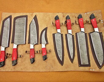 May include: Set of eight kitchen knives with red and black handles, arranged in a leather roll. The knives have a Damascus steel blade pattern.
