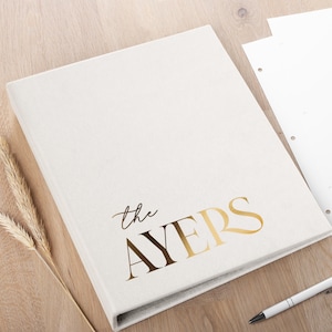 Könnte beinhalten: Ein cremefarbenes Fotoalbum mit dem Text "the AYERS" in goldfarbenen Buchstaben. Das Album liegt auf einer Holzoberfläche mit einem Stift, losem Papier und dekorativen Weizenähren.