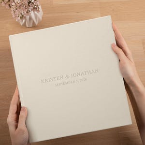 May include: A cream-colored photo album with the names "KRISTEN & JONATHAN" and the date "SEPTEMBER 5, 2026" embossed on the cover. The album is held open by two hands. A small vase of flowers sits in the background.