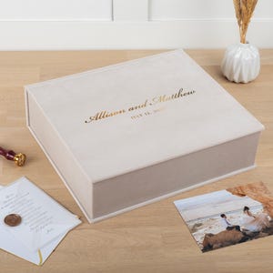 May include: A light beige, rectangular keepsake box with the names "Allison and Matthew" and the date "July 12, 2023" in gold lettering. A wedding photo and a card with a wax seal are next to the box. A small white vase with dried flowers is in the background.