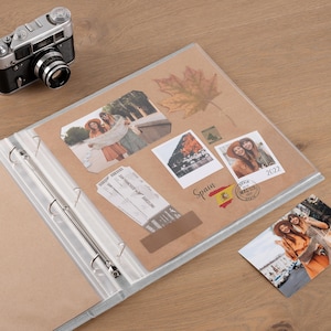 May include: A scrapbook with brown pages containing photographs, a dried maple leaf, a San Francisco to Barcelona airline ticket, and a Spain sticker with the text 2022. A vintage-style camera sits nearby, and an additional photograph lies loose.