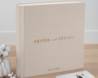May include: A beige linen-covered photo album with gold lettering that reads "AMANDA and EDWARD" and the date "07.12.2024". A small photo of a couple is on the wooden surface next to the album. The album is standing upright.