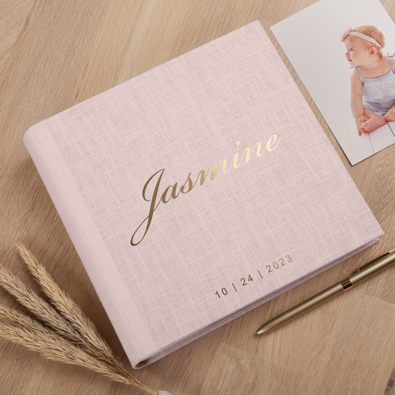 May include: A pale pink photo album with the name "Jasmine" in gold script. The date "10 | 24 | 2023" is printed below the name. A photo of a baby and a gold pen are next to the album.