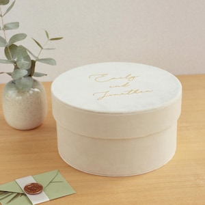 May include: A round, cream-colored velvet box with a lid featuring the gold-scripted text Emily and Jonathan. The box has a smooth, soft-touch finish and a cylindrical shape, presented as a decorative container for small items or gifts.