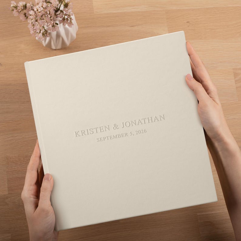 May include: A cream-colored photo album with the names "KRISTEN & JONATHAN" and the date "SEPTEMBER 5, 2026" embossed on the cover. The album is held open by two hands. A small vase of flowers sits in the background.