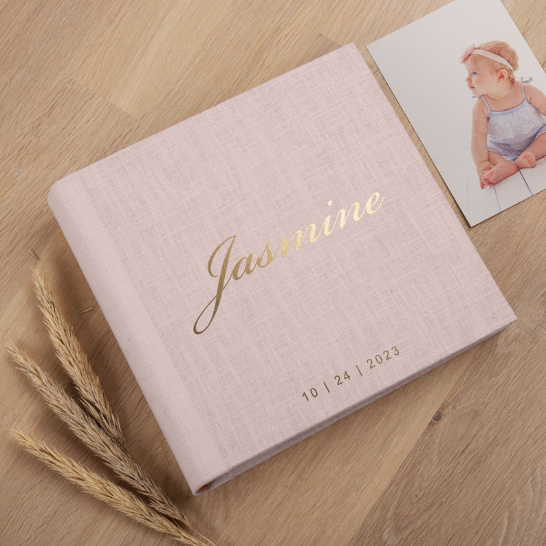 May include: A pale pink linen-effect photo album with the name "Jasmine" in gold script. The date "10 | 24 | 2023" is printed below the name. A photograph of a baby and dried wheat stalks are next to the album.