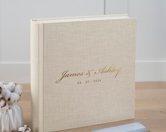 May include: A beige linen-textured photo album with gold lettering that reads "James & Ashley" and the date "04.27.2024". A printed photo of a couple on a beach is next to the album. The album is a wedding keepsake.