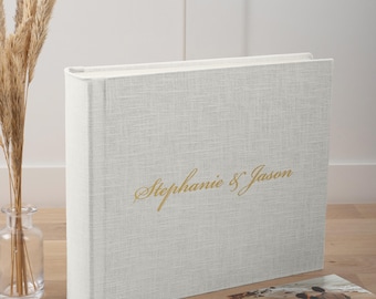 May include: A rectangular photo album with a light gray linen-textured cover. The front features the gold-colored cursive text Stephanie & Jason centered horizontally. The album is positioned upright with a visible spine and a textured fabric finish.