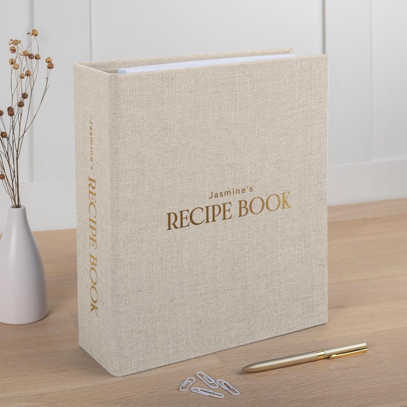 May include: A beige linen-textured recipe book with the text "Jasmine's RECIPE BOOK" in gold lettering. A gold pen and several paper clips are on the wooden surface next to the book. A white vase with dried flowers is in the background.