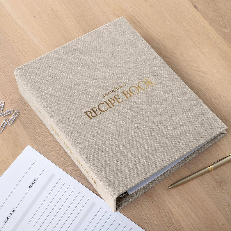 May include: A beige linen-covered recipe book with the words "Jasmine's RECIPE BOOK" in gold lettering. The book is open, revealing white pages. A gold pen and a sheet of paper with lines are next to the book.