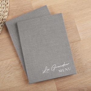 May include: Two rectangular menu covers featuring a textured grey fabric finish. The front cover displays the white printed text La Grandeur MENU in a combination of script and sans-serif fonts. The covers are stacked slightly offset from each other.