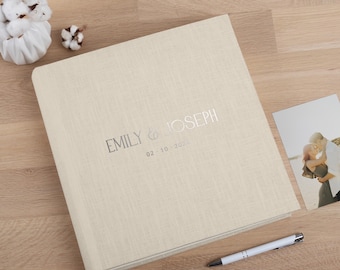May include: A beige linen-textured photo album with the names "EMILY & JOSEPH" in silver lettering and the date "02-10-2023". A photograph and a pen are next to the album. Decorative cotton flowers are in the background.