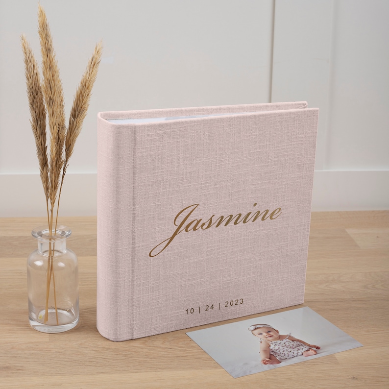 May include: A pale pink linen-effect photo album with the name "Jasmine" in gold script. The date "10 | 24 | 2023" is below the name. A small photo of a baby is next to the album. A clear glass vase with dried plants is on the left.