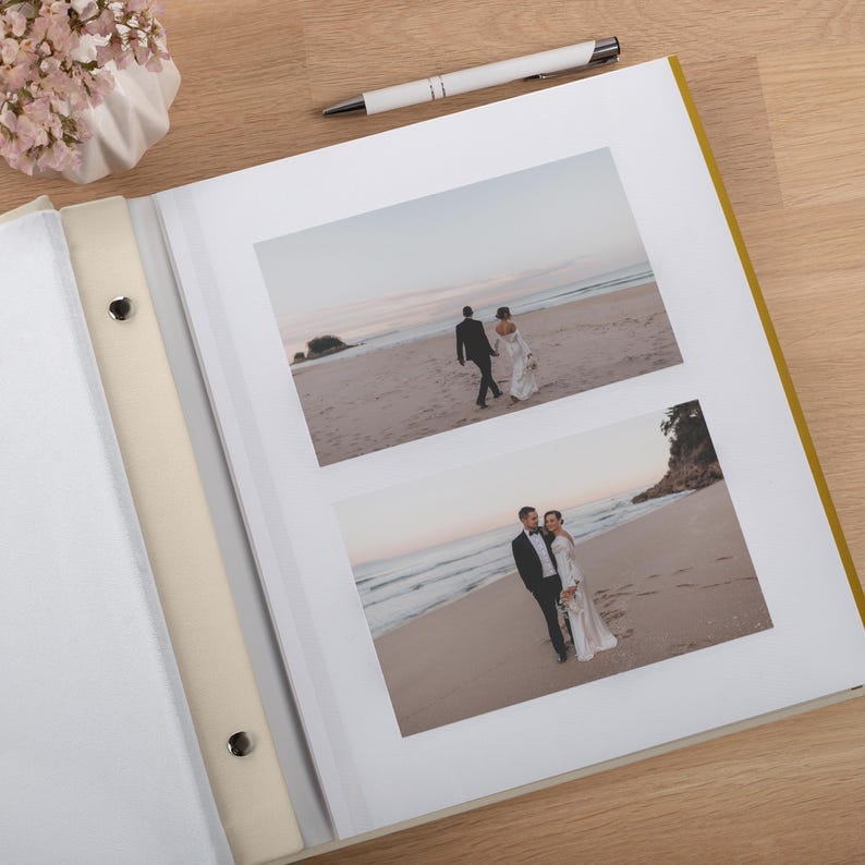 May include: A photo album with a cream-colored spine and metal screw posts, containing two landscape-oriented photographs of a couple on a beach. The top photo shows the couple walking away, and the bottom photo shows them standing together.