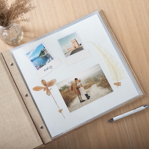 May include: An open photo album with a beige linen cover, displaying several photographs and decorative elements. The photos include travel scenes and a couple embracing. A pen and dried plants are also visible.