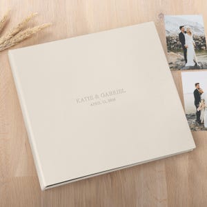 May include: A cream-colored wedding photo album with the names "KATIE & GABRIEL" and the date "APRIL 12, 2026" printed on the cover. Two wedding photos are visible on the right side of the album. The album is on a light wood surface.