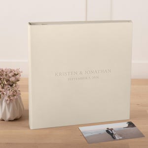 May include: A square, cream-colored photo album with a smooth cover featuring the embossed text KRISTEN & JONATHAN and SEPTEMBER 5, 2026. A small rectangular photograph of a couple on a beach is placed in front of the album on a wooden surface.
