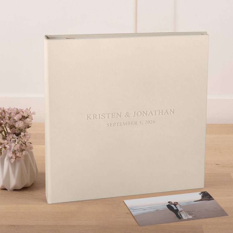 May include: A square, cream-colored photo album with a smooth cover featuring the embossed text KRISTEN & JONATHAN and SEPTEMBER 5, 2026. A small rectangular photograph of a couple on a beach is placed in front of the album on a wooden surface.