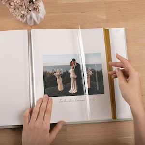 May include: A photo album with white pages and a gold spine, featuring a photograph of a couple in wedding attire standing outdoors near the ocean. The text "Two hearts in love need no words" is printed below the photo, which is covered by a clear film.