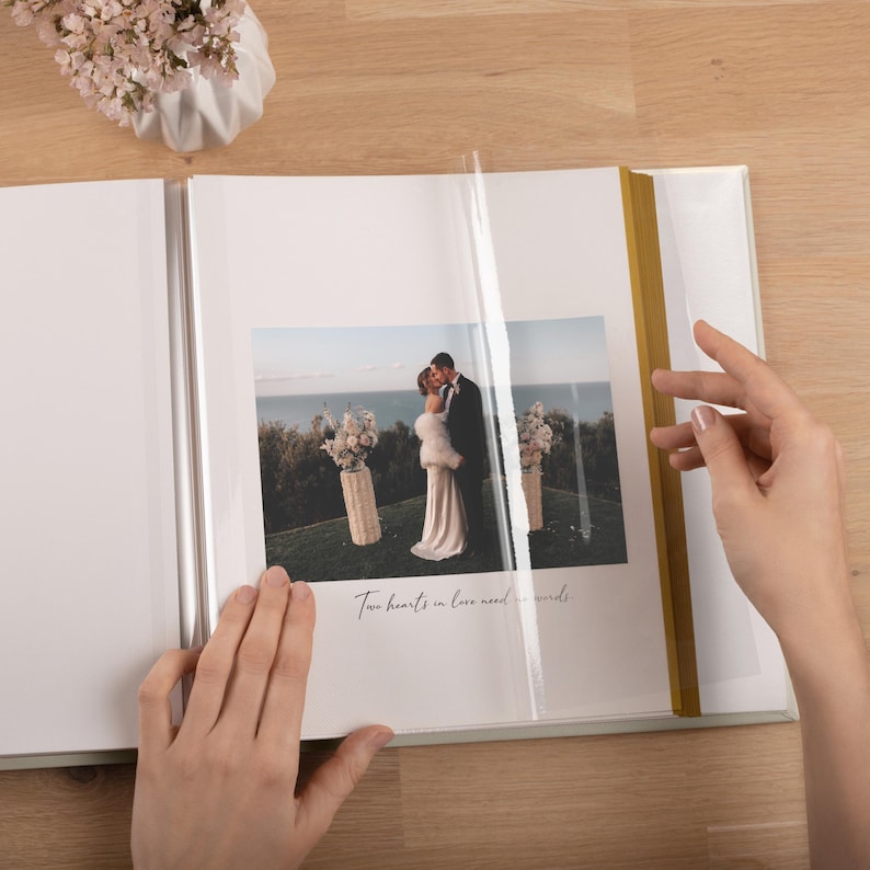 May include: A photo album with white pages and a gold spine, featuring a photograph of a couple in wedding attire standing outdoors near the ocean. The text "Two hearts in love need no words" is printed below the photo, which is covered by a clear film.