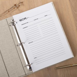 May include: A recipe binder with a linen cover, open to a blank recipe page. The page has lines for recipe details, including preparation time, ingredients, directions, and notes. A gold pen rests on the wooden surface.