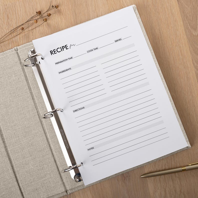 May include: A recipe binder with a linen cover, open to a blank recipe page. The page has lines for recipe details, including preparation time, ingredients, directions, and notes. A gold pen rests on the wooden surface.